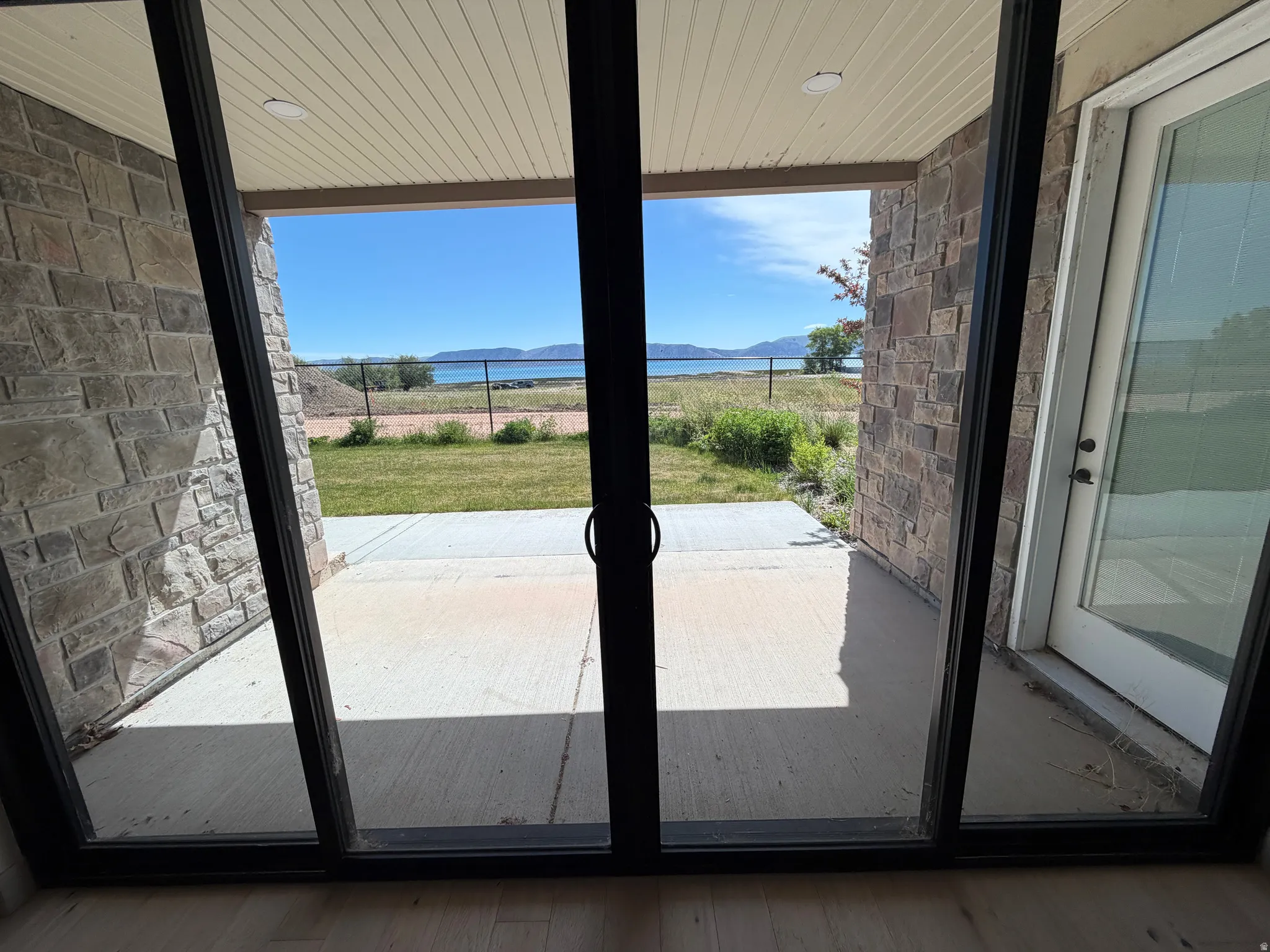 Sliding glass door opening to a covered patio with stone facade, concrete flooring, and a serene water backdrop framed by distant mountains