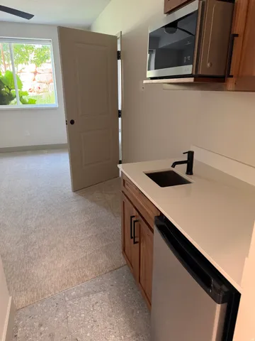 Wet bar featuring a stainless steel microwave, wood-finish cabinetry with black hardware, white countertop with an undermount sink and matte black faucet, and a stainless steel compact refrigerator