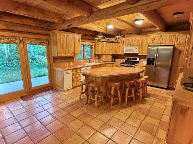 Kitchen featuring exposed log beams, extensive wood cabinetry, a central island with integrated seating, stainless steel appliances, and terra cotta floor tiles
