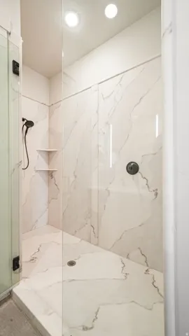 Walk-in shower with full-height white marble-look tiling, glass enclosure, recessed lighting, built-in corner shelves, and a matte black shower head and controls