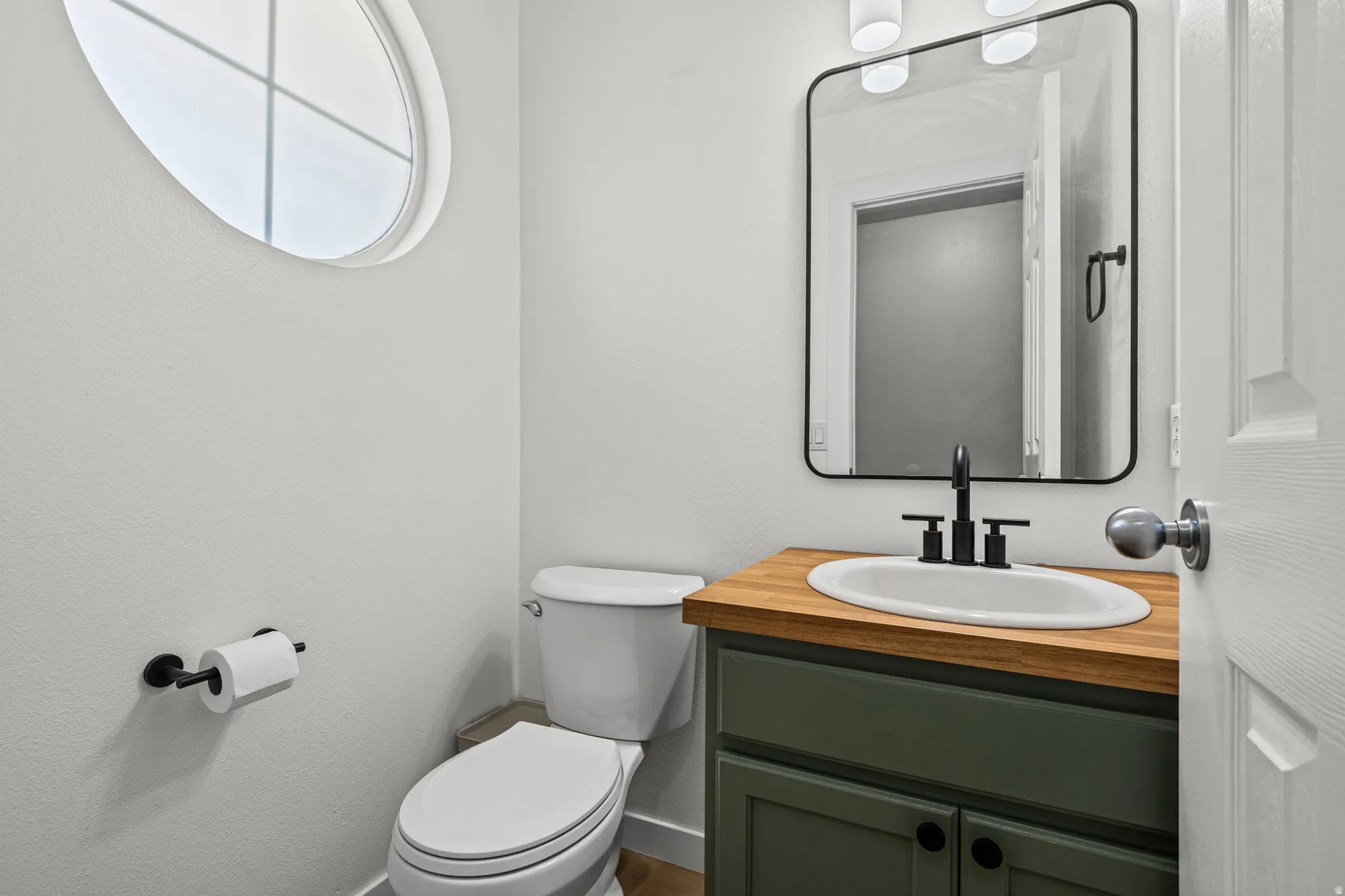 Powder room featuring a circular window, a vanity with a wood-finish countertop, a sage green cabinet, a rectangular mirror, and matte black fixtures