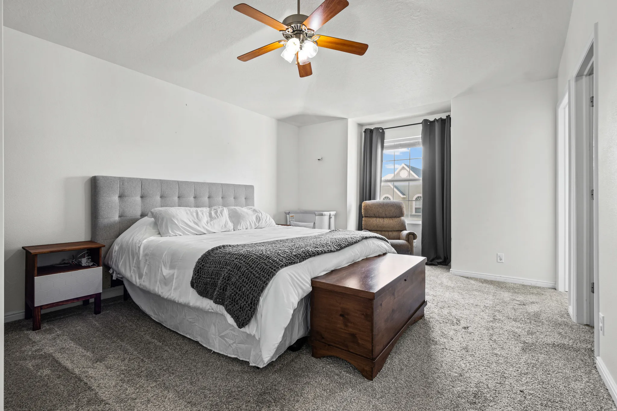 Spacious room featuring neutral carpeting, light-colored walls, a ceiling fan with light fixture, and a window with dark drapery