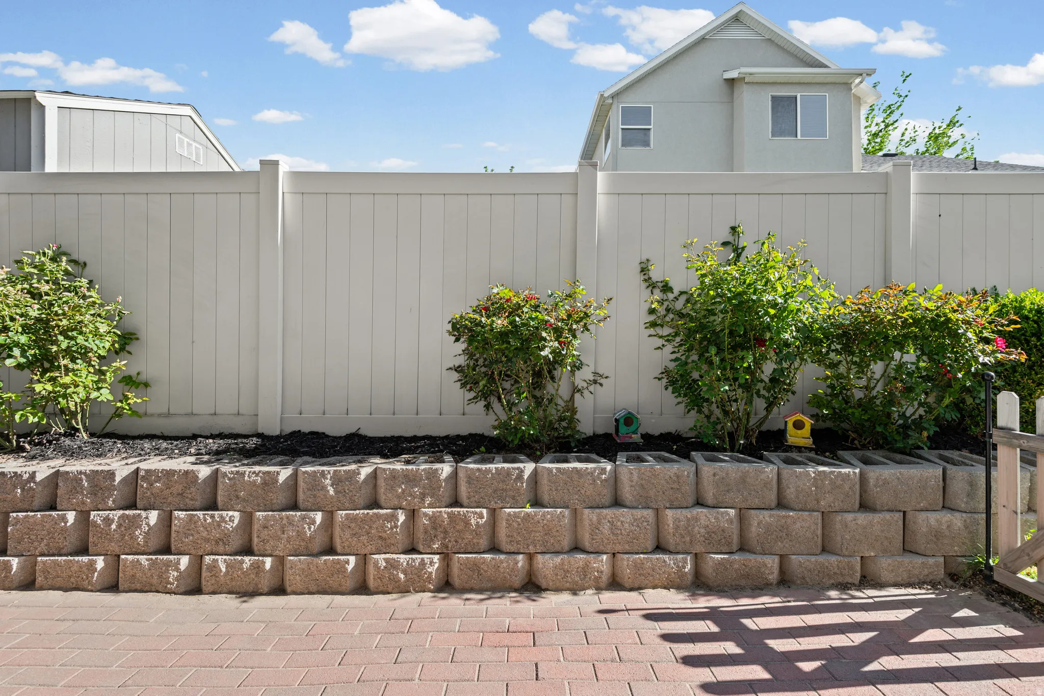 Brick paver patio featuring a stacked block retaining wall, mature shrubs, and a white privacy fence