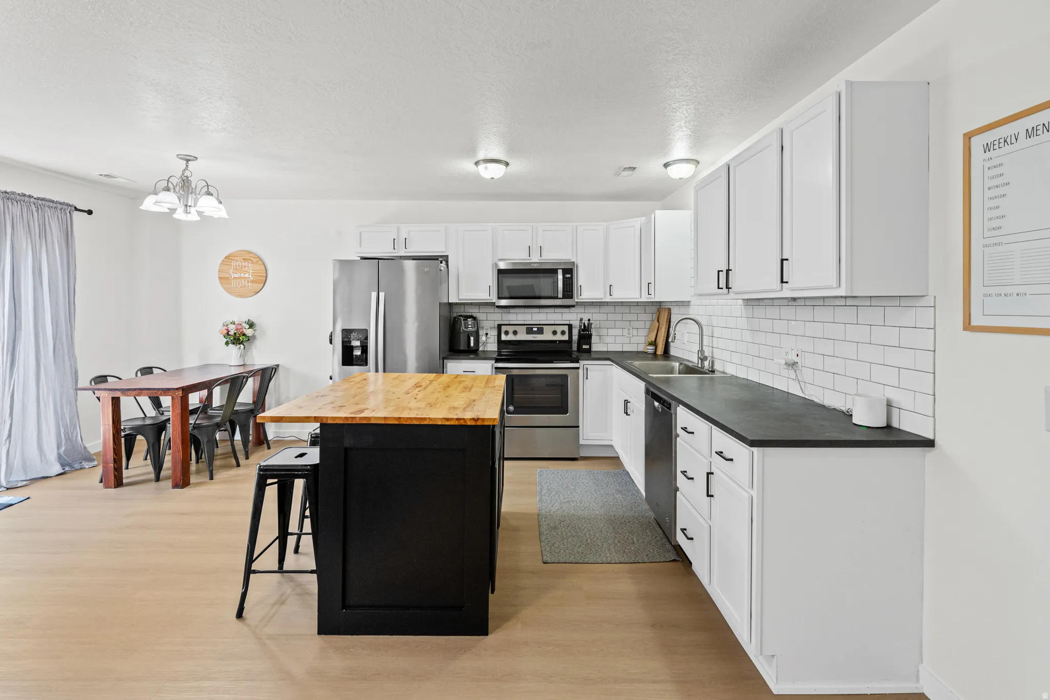 Contemporary kitchen featuring a prominent island with a wood countertop, white cabinetry with black hardware, dark countertops, stainless steel appliances, and a white subway tile backsplash