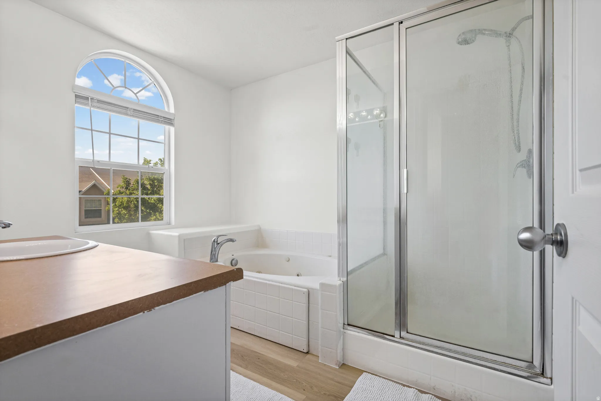 Bathroom featuring a large arched window, a built-in jetted tub with white tile surround, a separate glass-enclosed shower, a vanity with an integrated sink, and wood-finish flooring