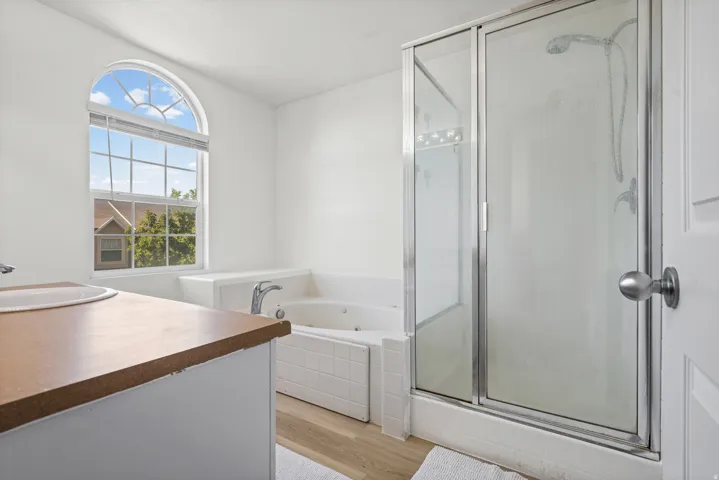 Bathroom featuring a large arched window, a built-in jetted tub with white tile surround, a separate glass-enclosed shower, a vanity with an integrated sink, and wood-finish flooring