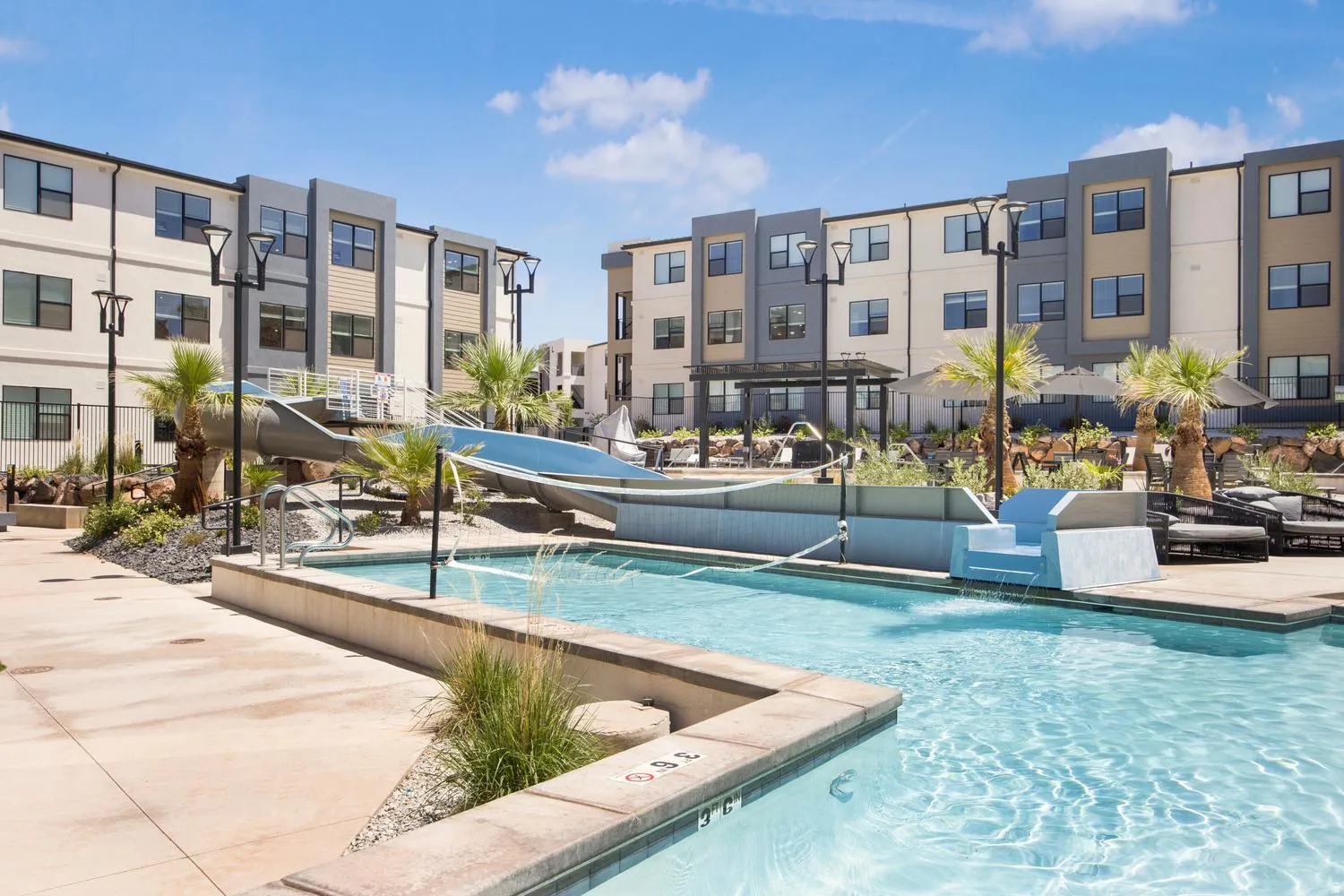 Resort-style pool area featuring multiple swimming zones, an integrated water slide, contemporary lounge seating, and lush palm trees