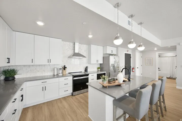 Modern kitchen featuring an expansive island with seating, pendant lighting, stainless steel appliances, white cabinetry, and a chevron tile backsplash