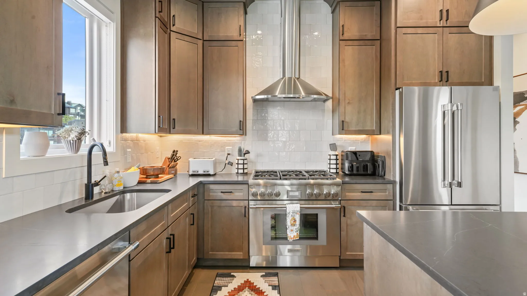 Contemporary kitchen featuring stainless steel appliances, a gas range with an overhead vent hood, ample counter space, a double-basin sink with a matte black faucet, and wood-finish cabinetry