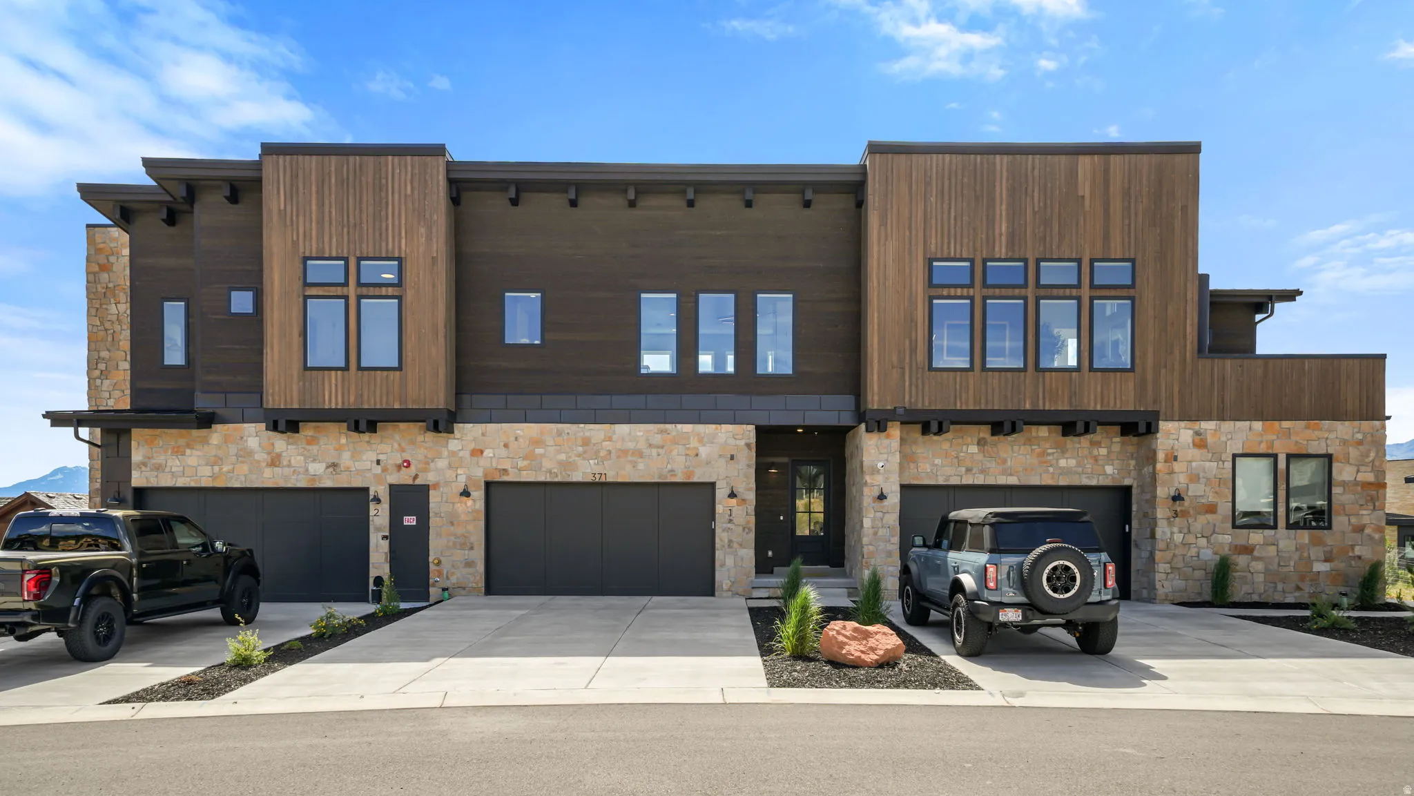 Contemporary multi-level facade featuring stone and wood paneling, multiple garages with dark doors, large black-framed windows, and a paved driveway