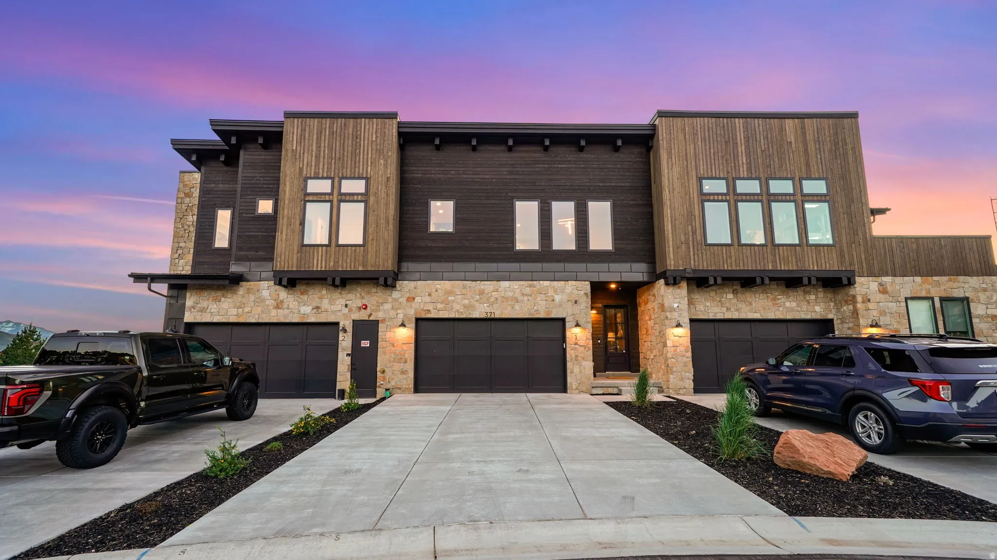 Modern facade featuring stone veneer, vertical wood paneling, dark horizontal siding, multiple garages with dark doors, and concrete driveways