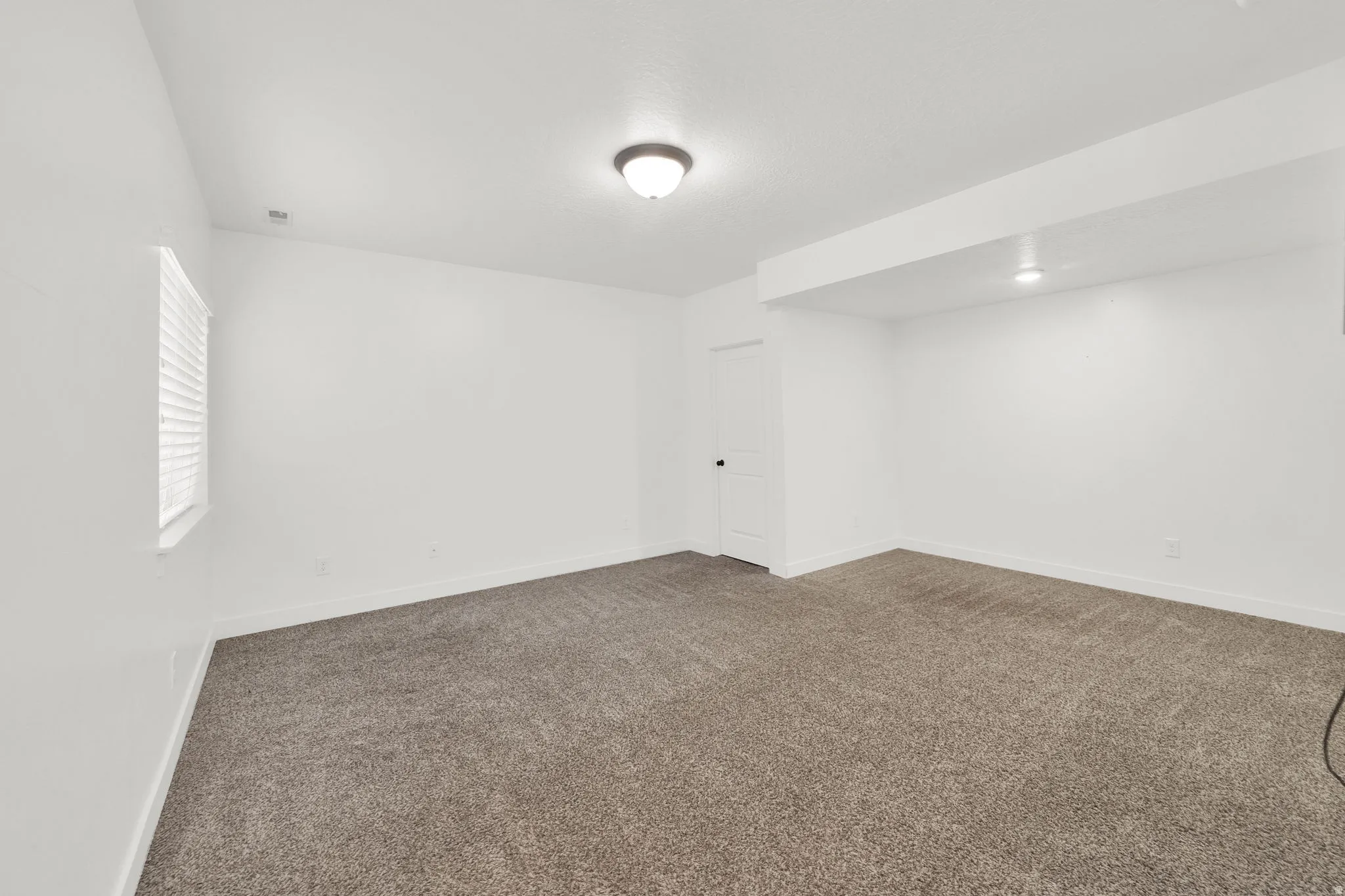 Spacious carpeted room featuring a single window with blinds, ceiling-mounted light fixture, recessed lighting, and white walls