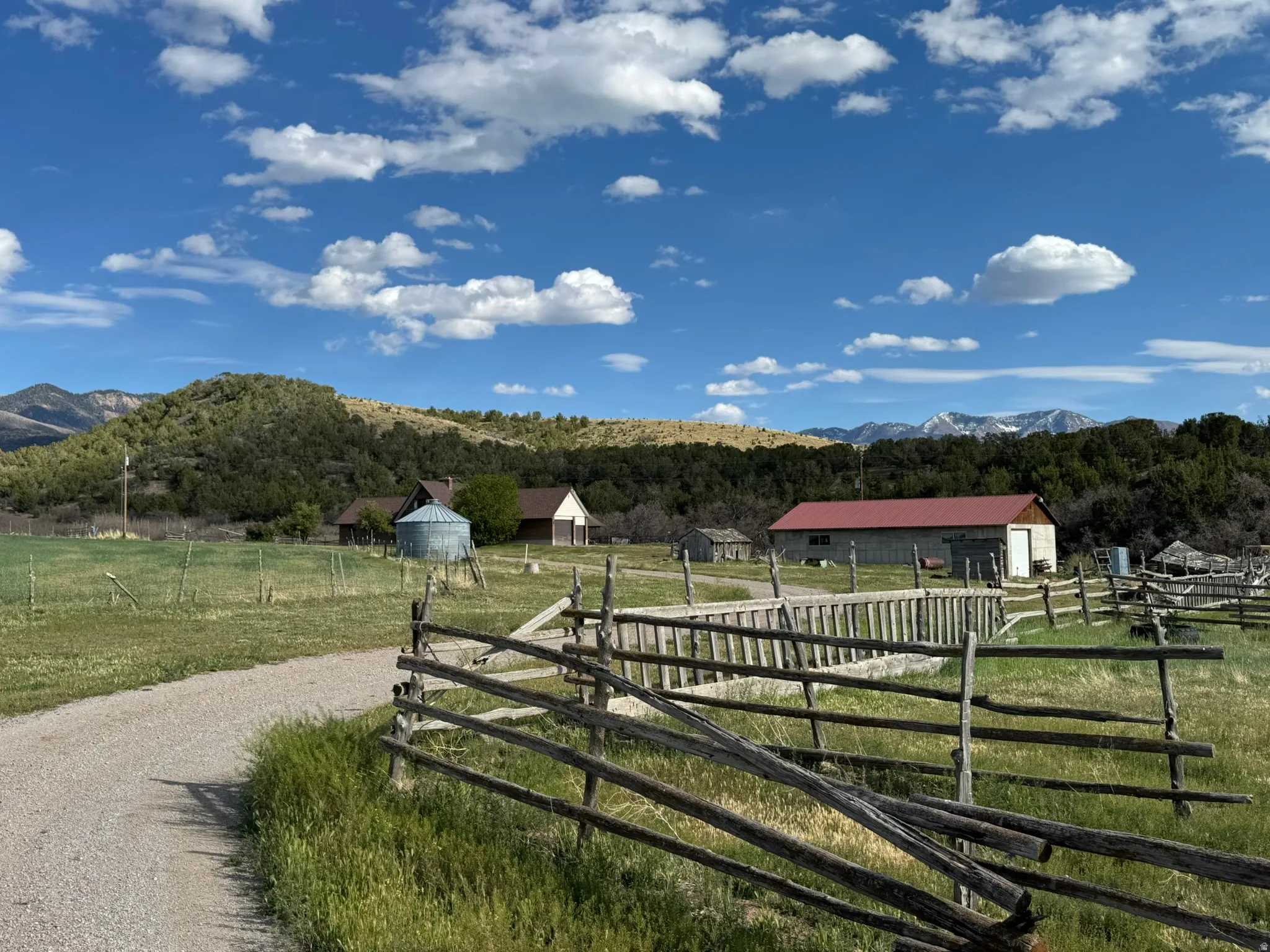 Expansive rural property featuring a gravel driveway, split-rail fencing, multiple outbuildings, and distant mountain backdrops