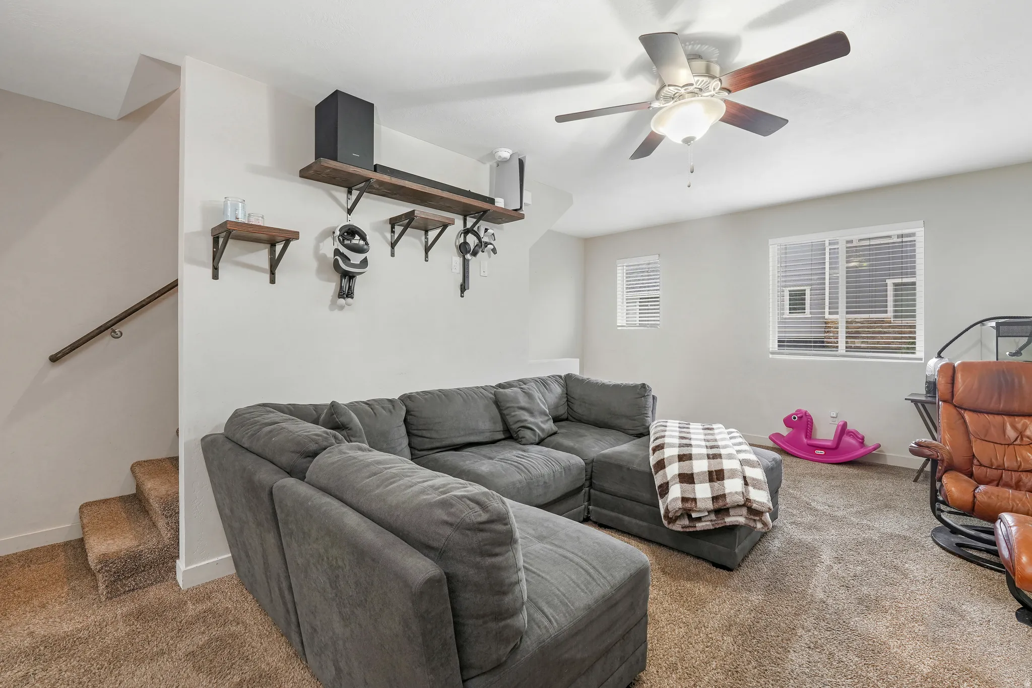 Carpeted living area featuring a ceiling fan with integrated lighting, wood-finish shelving, and multiple windows with blinds