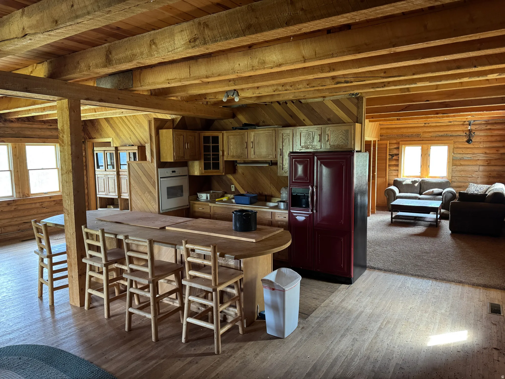 Open-concept living space featuring wood-finish flooring, exposed wood beam ceiling, wood-paneled walls, a kitchen island with seating, and built-in oven