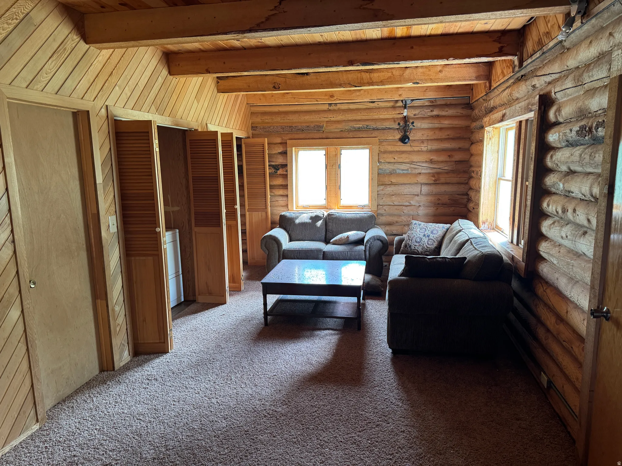 Living space featuring exposed log walls, a vaulted wood-paneled ceiling with exposed beams, and light-toned carpeting