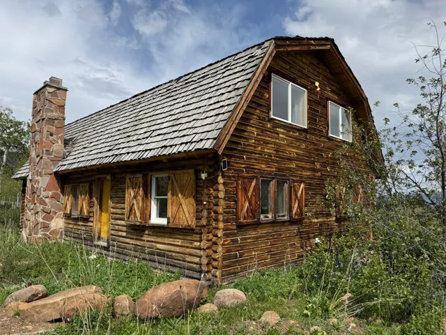 Rustic log cabin exterior featuring a cedar shake roof, stone chimney, wood-clad siding, and custom wood window shutters