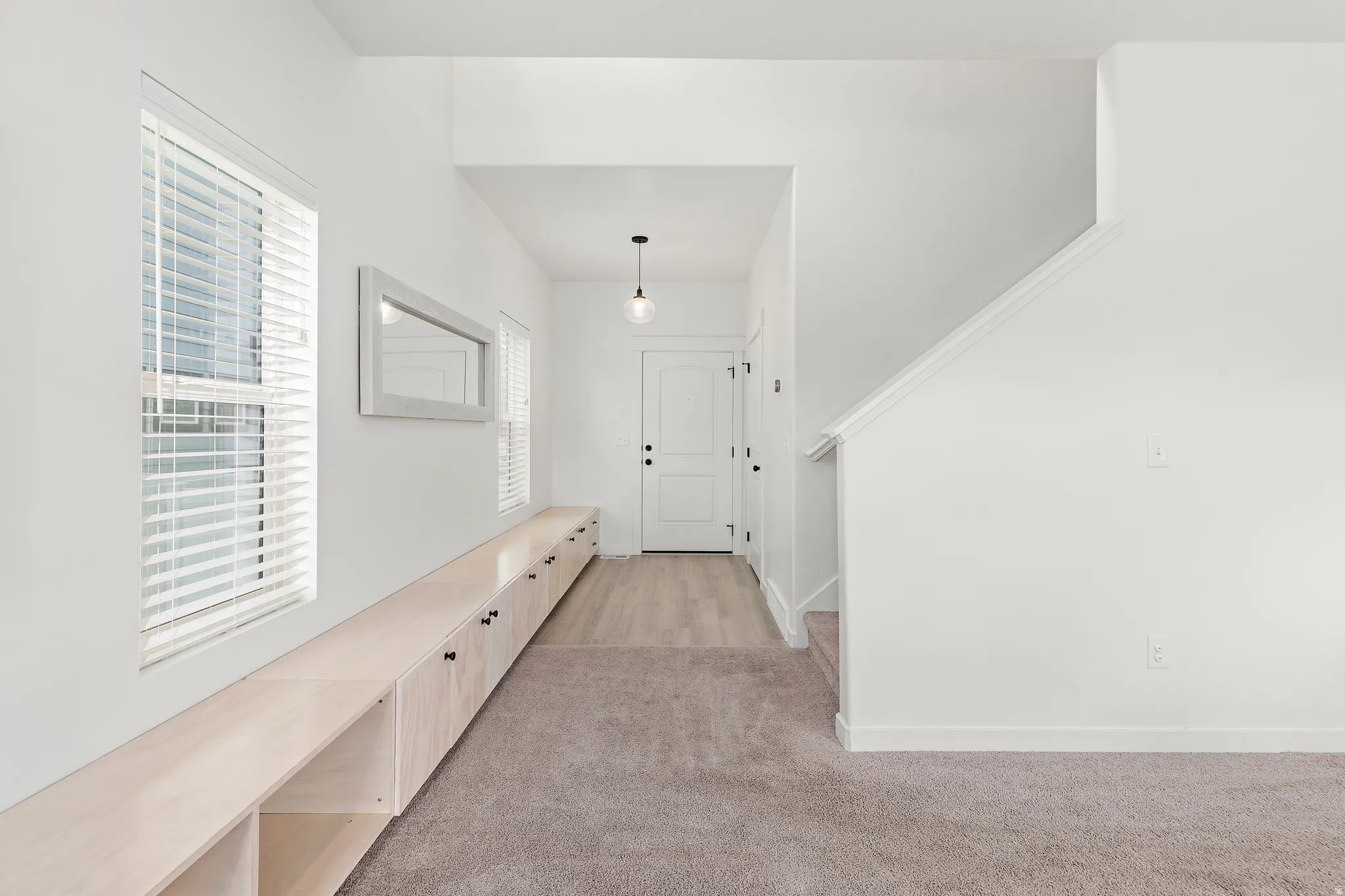 Bright entryway featuring a natural wood-finish storage bench with open shelving, two windows with white blinds, and a staircase with white trim