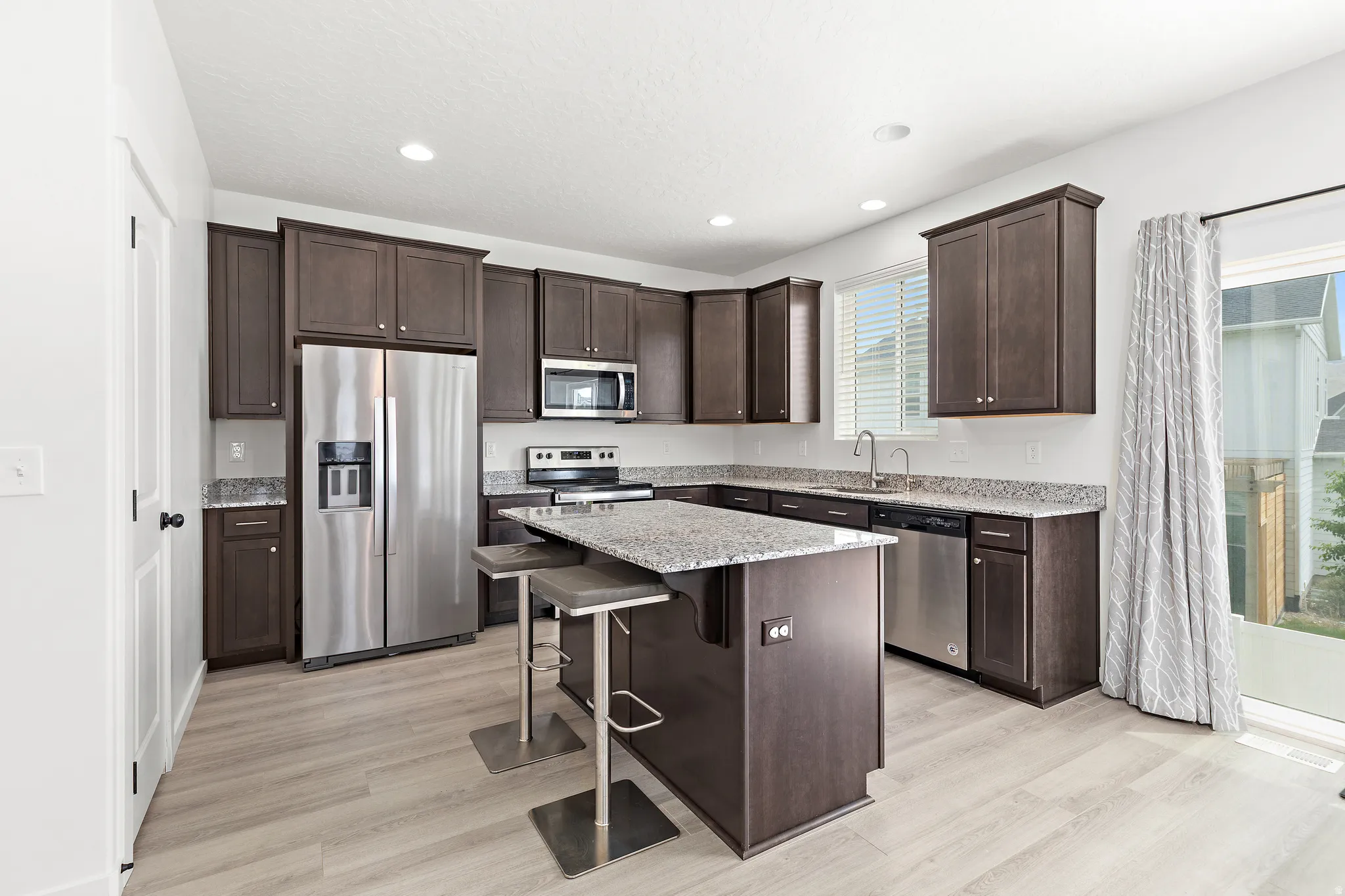 Modern kitchen featuring stainless steel appliances, dark wood cabinetry, light-toned wood-finish flooring, granite countertops, and a sliding glass door