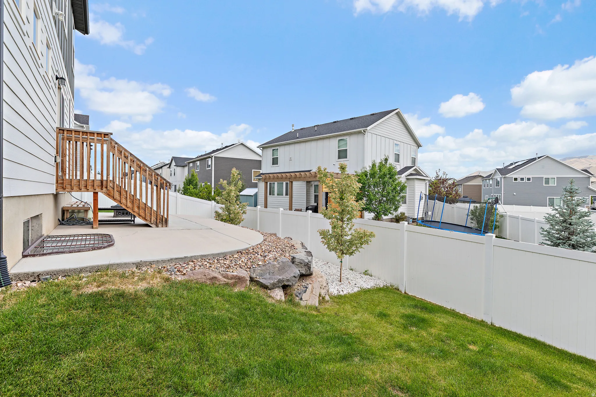 Rear exterior featuring a concrete patio, wood deck with stairs, white vinyl fencing, and a sloped lawn