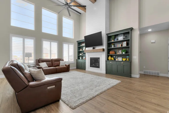 Vaulted living space featuring multiple tall windows with horizontal blinds, wood-finish flooring, a contemporary ceiling fan, and a fireplace with a shiplap surround and wood mantel