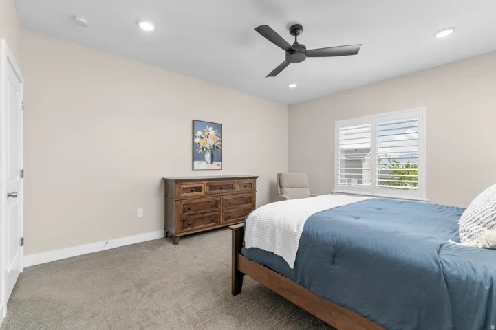 Spacious bedroom featuring neutral wall tones, recessed lighting, a ceiling fan with matte black blades, and a window with white plantation shutters