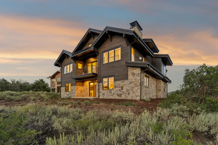 Multi-level residence featuring a dark wood-finish exterior, stone accents, multiple large windows, a second-story balcony with metal railing, and a prominent chimney structure