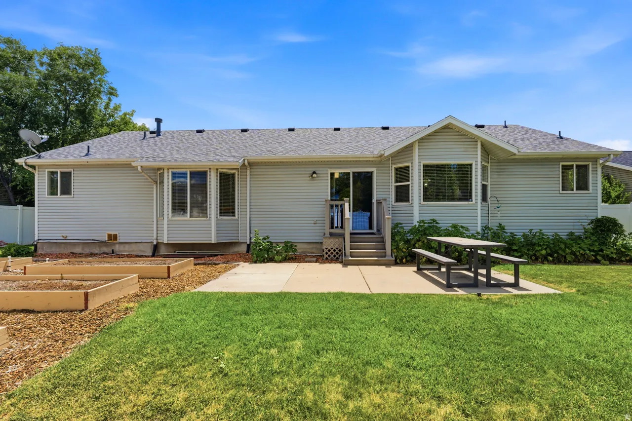 Rear exterior with light gray siding, featuring a concrete patio, a sliding glass door with wood steps, a bay window, and several raised garden beds