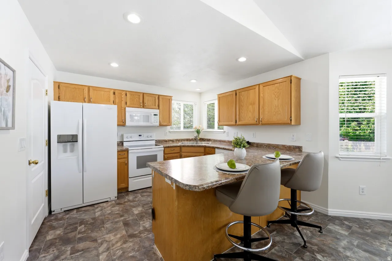 Kitchen featuring wood cabinetry, white appliances, a peninsula island with counter seating, recessed lighting, and dual windows