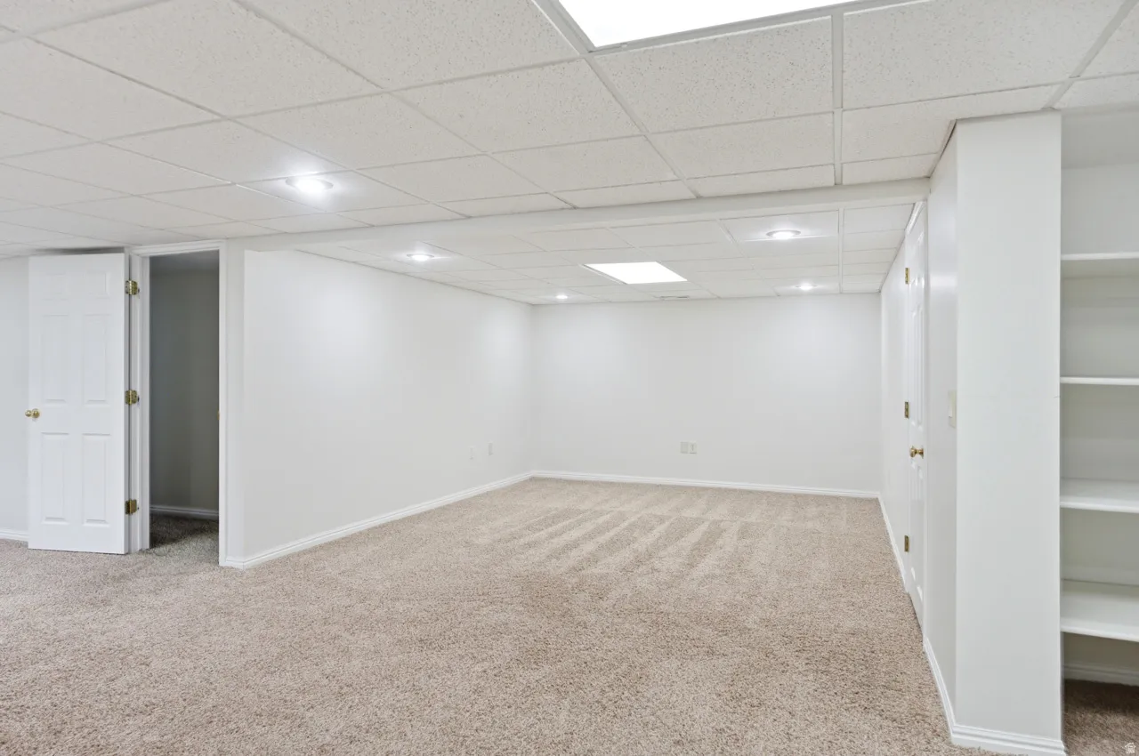 Spacious room featuring neutral carpeting, a drop-tile ceiling with recessed lighting and a flush-mount fixture, built-in shelving, and white interior doors
