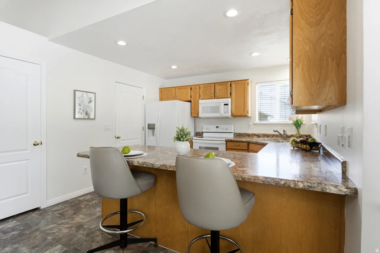 Kitchen featuring a breakfast bar with stone-finish countertops, wood cabinetry, white appliances, recessed lighting, and resilient flooring