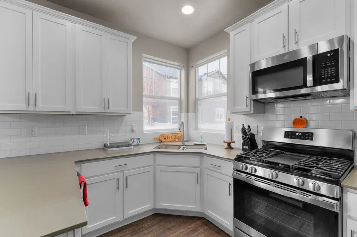 White shaker cabinetry with brushed nickel hardware, stainless steel appliances, corner sink with dual windows, subway tile backsplash, and wood-finish flooring