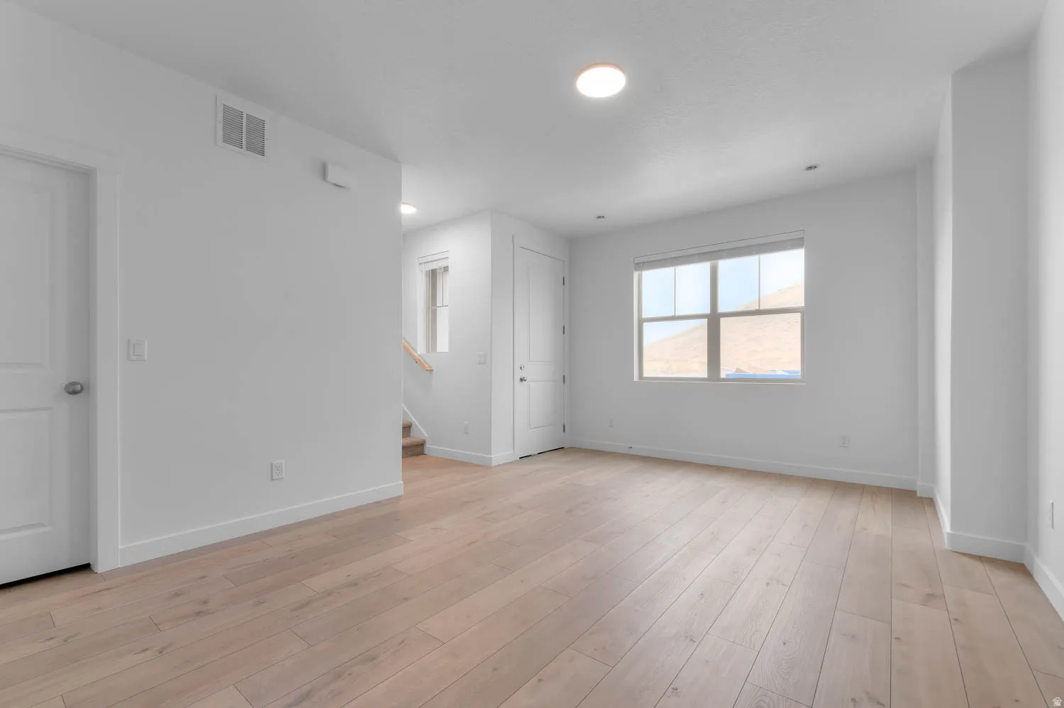 Spacious room featuring light wood-finish flooring, white walls, and a large window with a roller shade