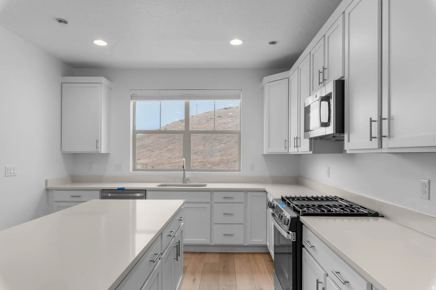 Open-concept kitchen featuring an expansive island, light-toned countertops, white shaker cabinetry, stainless steel appliances, and wood-finish flooring