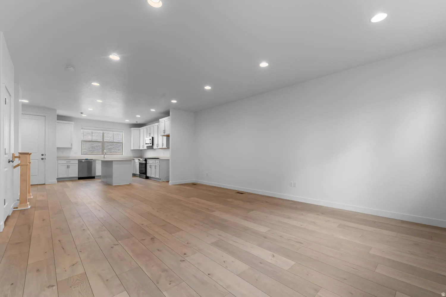 Open-concept interior featuring light wood-finish flooring, recessed lighting, and a white kitchen with an island, stainless steel appliances, and white cabinetry