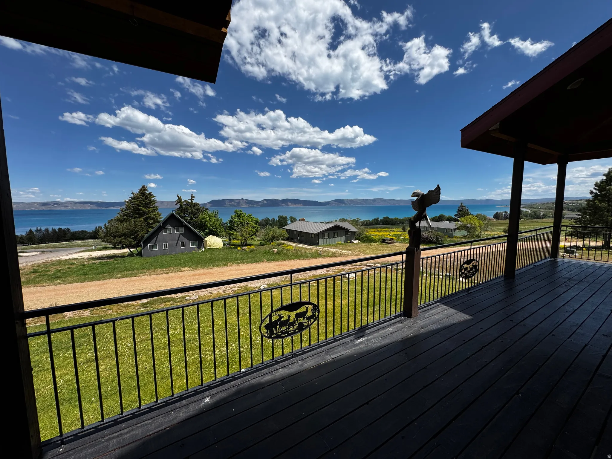 Expansive outdoor deck with dark wood-finish flooring and black metal railing, featuring a lake and distant hills backdrop