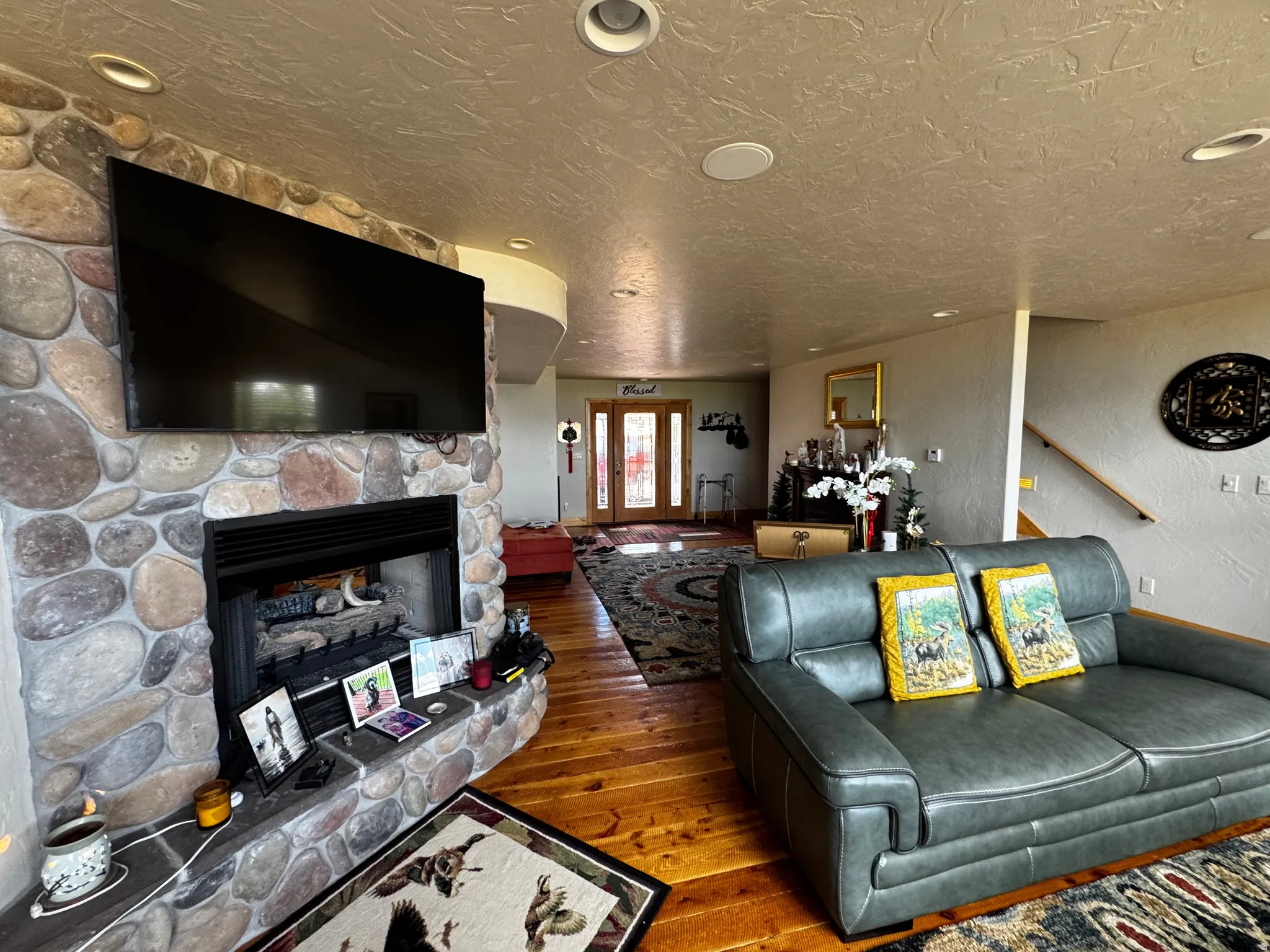 Stone-clad fireplace with a hearth and integrated television mount, complemented by wood-finish flooring, recessed lighting, and a partial view of a staircase with a wooden handrail