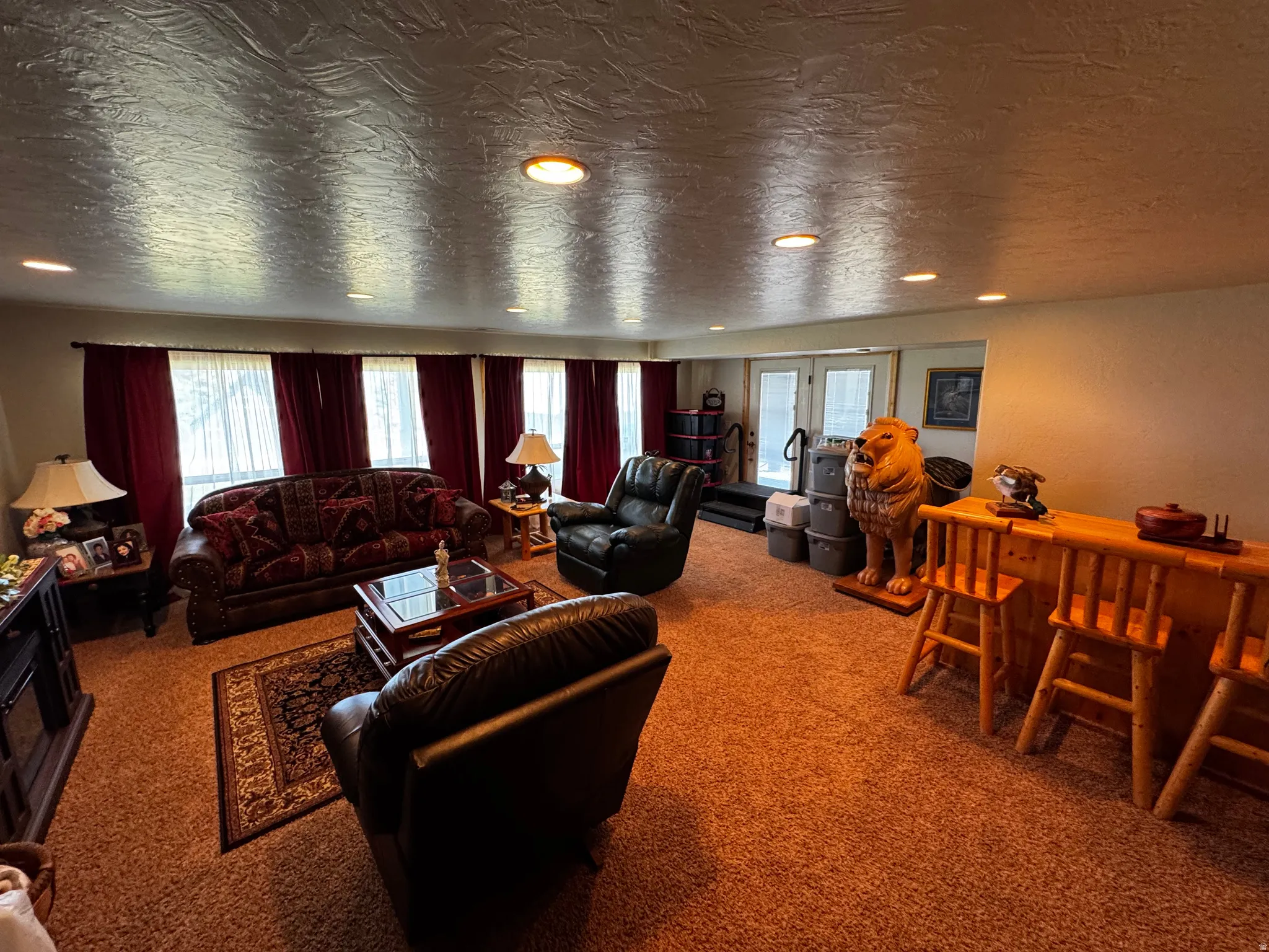 Recessed lighting illuminates a spacious room featuring a wood-finish bar with two stools, light-toned carpeting, and multiple windows with red curtains