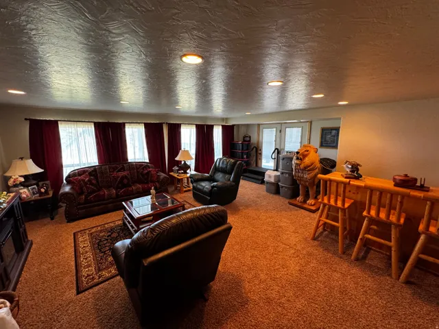 Recessed lighting illuminates a spacious room featuring a wood-finish bar with two stools, light-toned carpeting, and multiple windows with red curtains