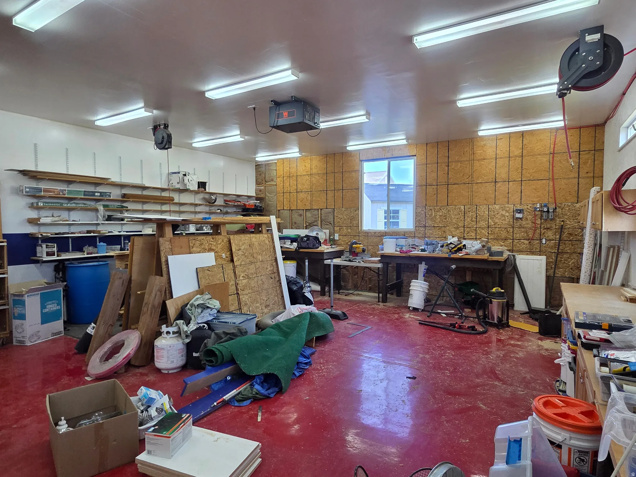 Spacious workspace featuring a red-finished concrete floor, multiple fluorescent light fixtures, built-in shelving, and a wall-mounted air hose reel