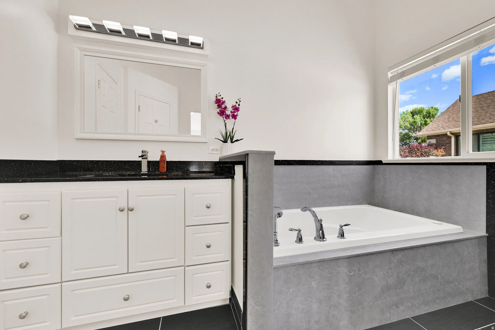 Built-in soaking tub with chrome fixtures, white vanity with black countertop, and a large mirror with overhead lighting