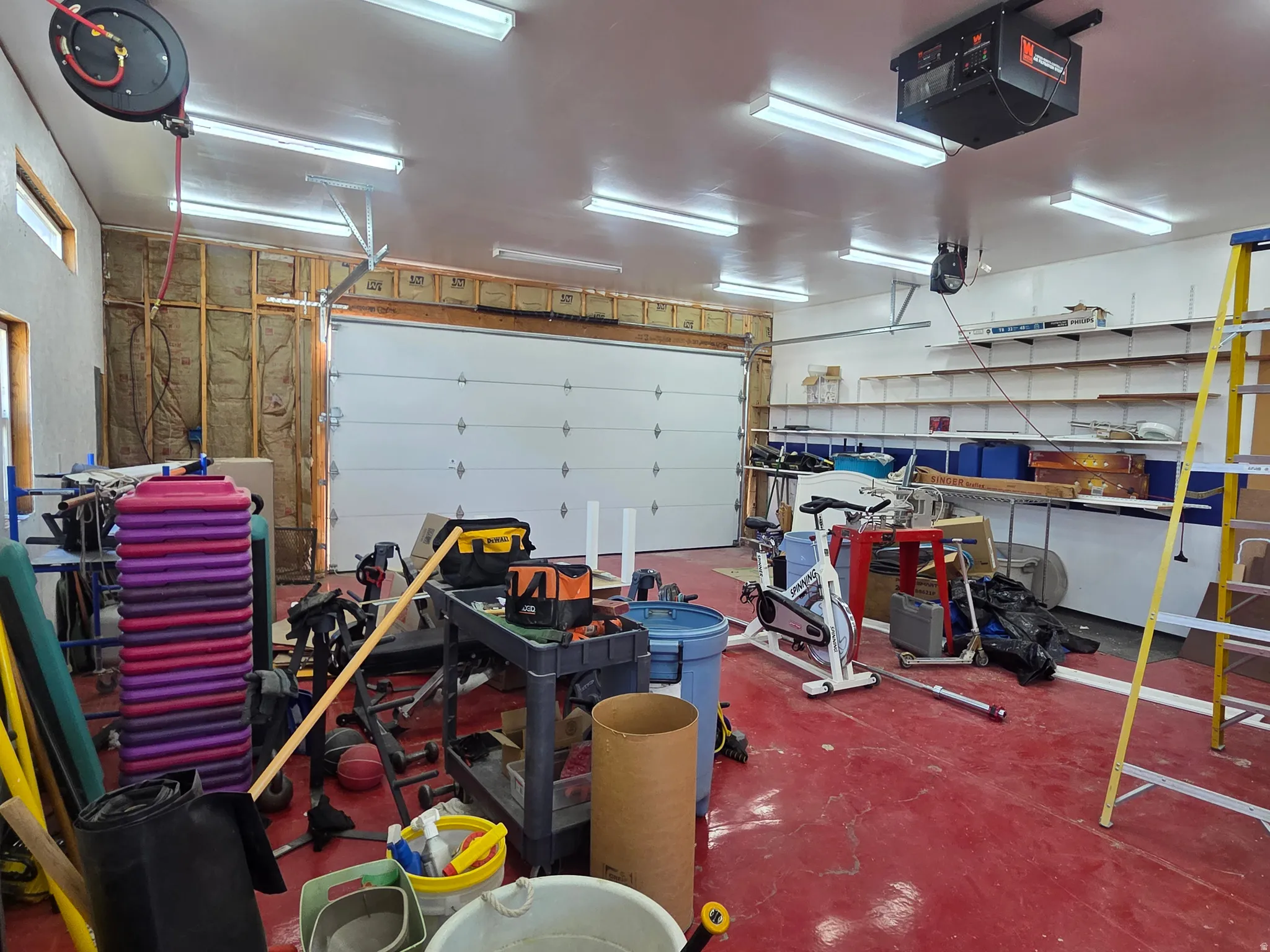 Spacious garage featuring a sectional garage door, multiple fluorescent ceiling lights, built-in wall shelving, and a red-painted concrete floor