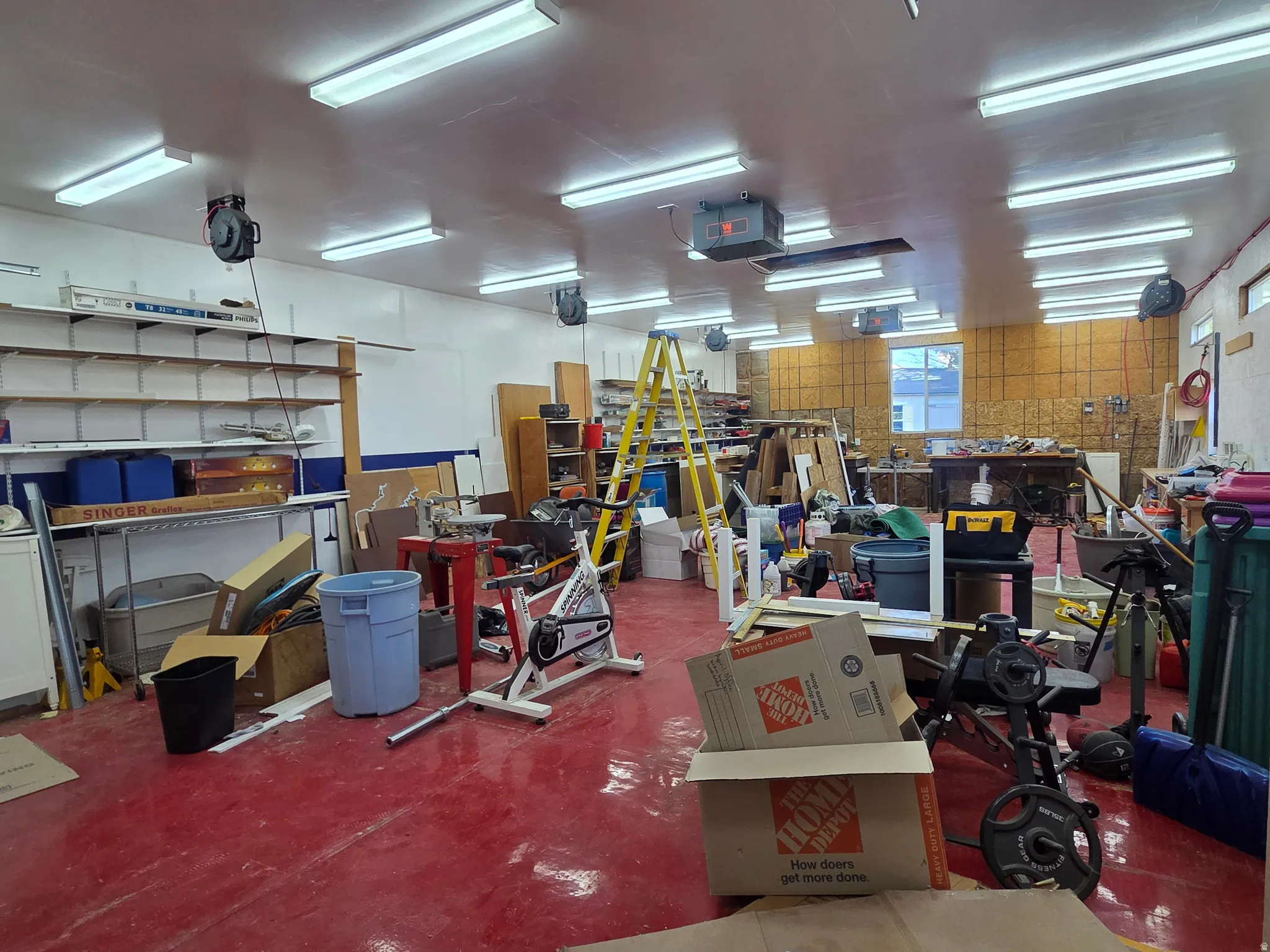 Spacious interior workshop area featuring a red finished concrete floor, extensive fluorescent lighting, multiple electrical outlets, built-in shelving, and a large window providing natural light