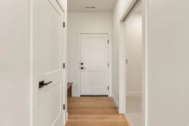 Interior hallway featuring light wood-finish flooring, white paneled doors with matte black hardware, and a recessed overhead vent