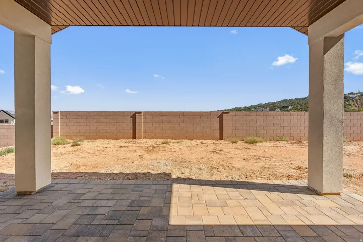 Covered patio with decorative pavers and concrete support columns, opening to a spacious dirt lot with brick perimeter fencing