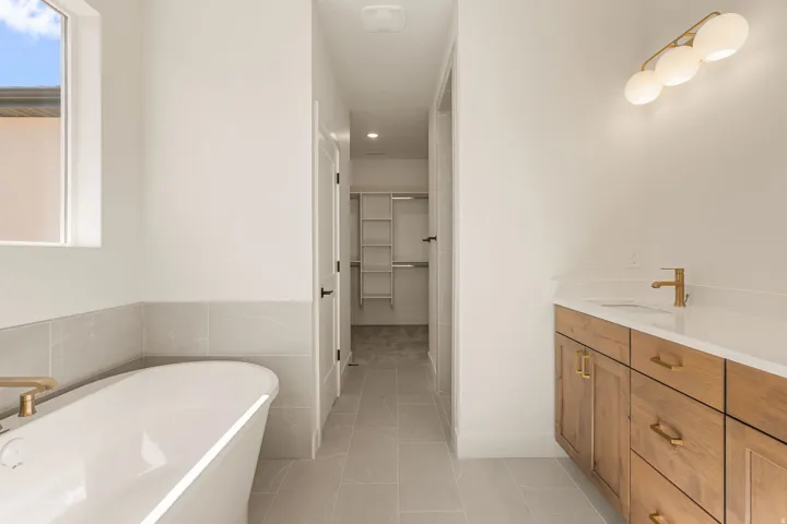 Bathroom featuring a freestanding soaking tub with a gold-finish faucet, a window with natural light, a wood-finish vanity with a white countertop and undermount sink, and light gray floor tiling