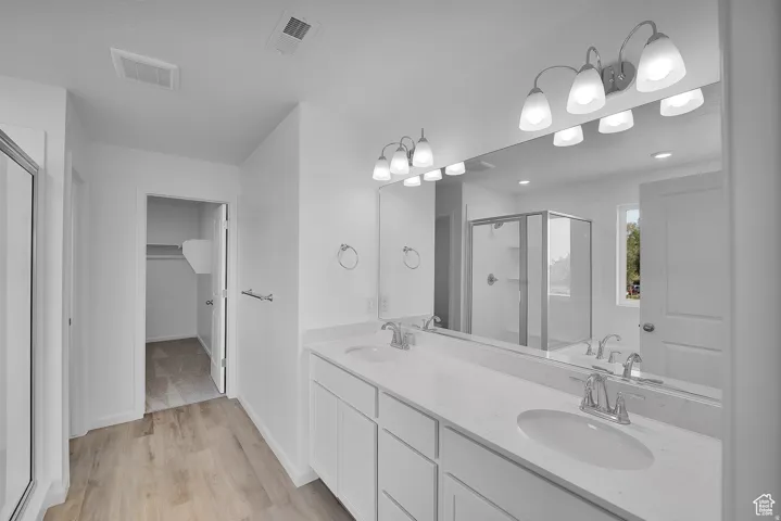 Bathroom featuring a dual vanity with undermount sinks and light-toned countertops, wood-finish flooring, a glass-enclosed shower, and a large mirror with overhead lighting