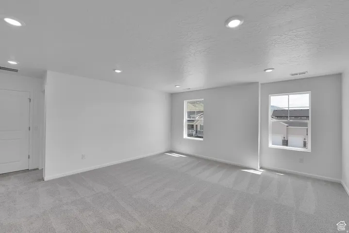 Spacious room featuring light gray carpeting, neutral wall finishes, multiple recessed ceiling lights, and two large windows with white trim