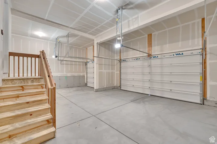 Spacious garage featuring a concrete floor, two white panel garage doors, integrated overhead lighting, exposed wood framing, and a natural wood staircase