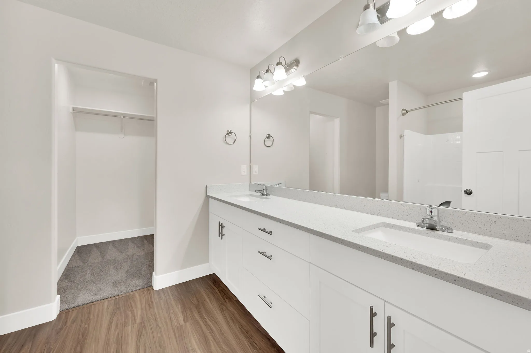 Dual vanity with a stone-finish countertop, wood-finish flooring, full-width mirror, and an adjacent walk-in closet with shelving